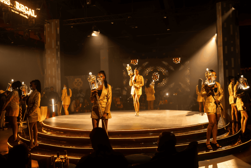 A luxurious experiential marketing activation featuring brand ambassadors in gold outfits presenting illuminated Chivas Regal bottles on a stage, engaging the audience in a high-end nightclub setting.